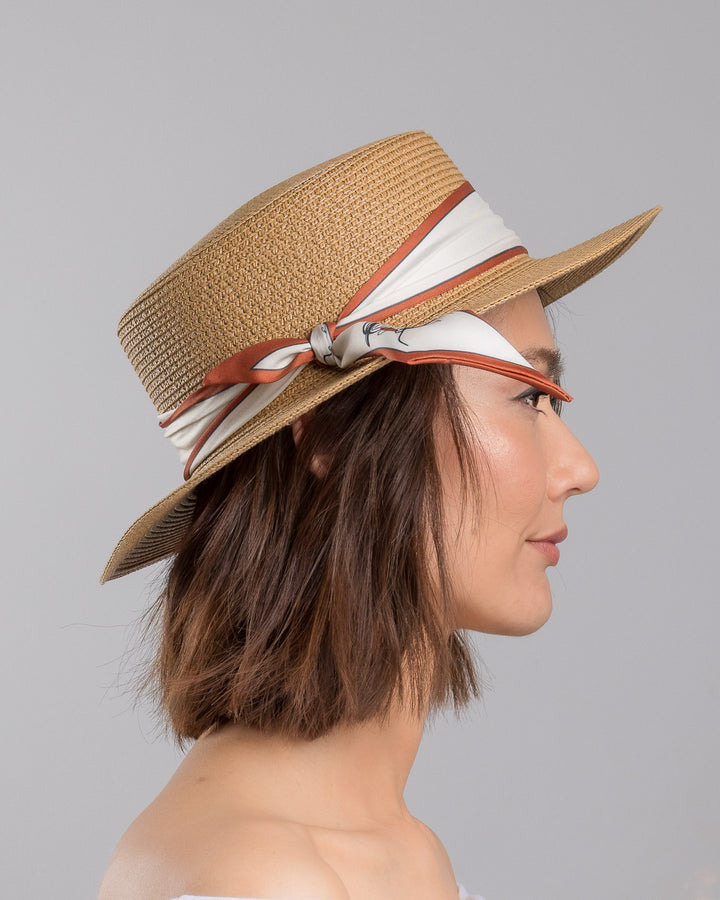 The model is showcasing the right side close up of the natural woven straw boater hat with a wide brim, wrapped in a cream and rust silk ribbon and finished with a tied scarf featuring an illustrated female portrait detail. Design and made by Merve Bayindir millinery.