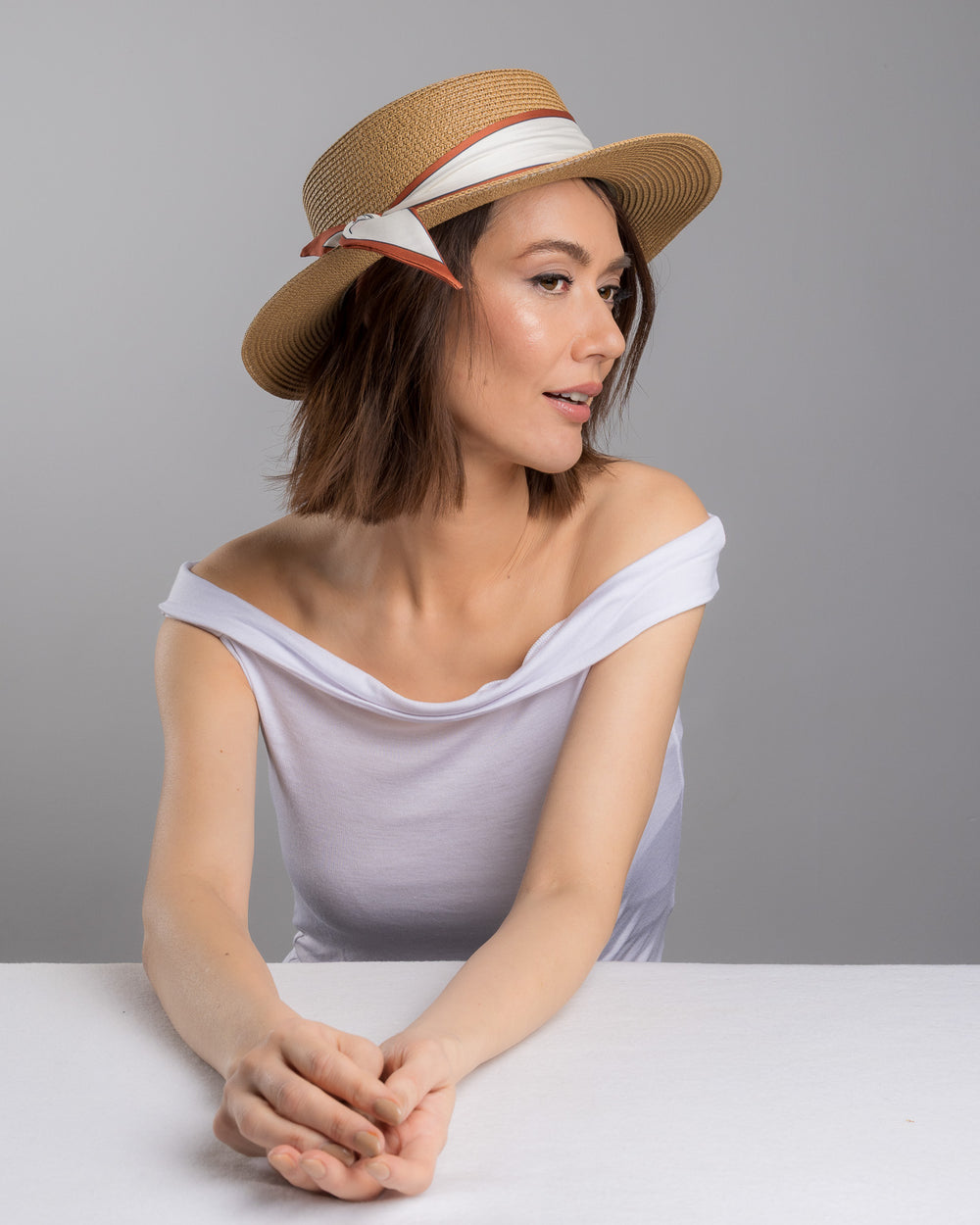 The female model is posing, wearing Tarin, the natural woven straw boater hat with a wide brim, wrapped in a cream and rust silk ribbon and finished with a tied scarf featuring an illustrated female portrait detail. Design and made by Merve Bayindir millinery.