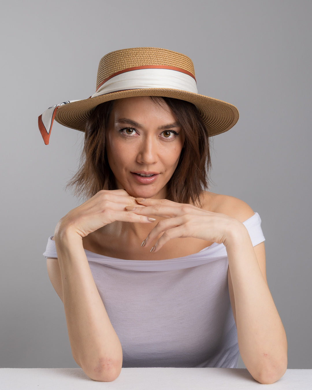 The female model is posing, wearing Tarin, the natural woven straw boater hat with a wide brim, wrapped in a cream and rust silk ribbon and finished with a tied scarf featuring an illustrated female portrait detail. Design and made by Merve Bayindir millinery.