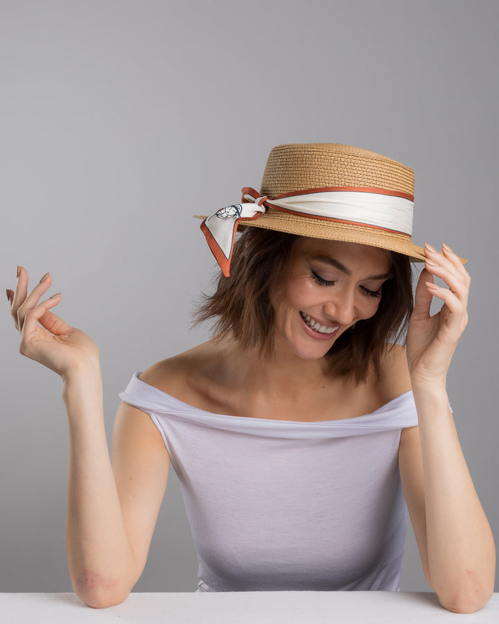The female model is posing, wearing Tarin, the natural woven straw boater hat with a wide brim, wrapped in a cream and rust silk ribbon and finished with a tied scarf featuring an illustrated female portrait detail. Design and made by Merve Bayindir millinery.
