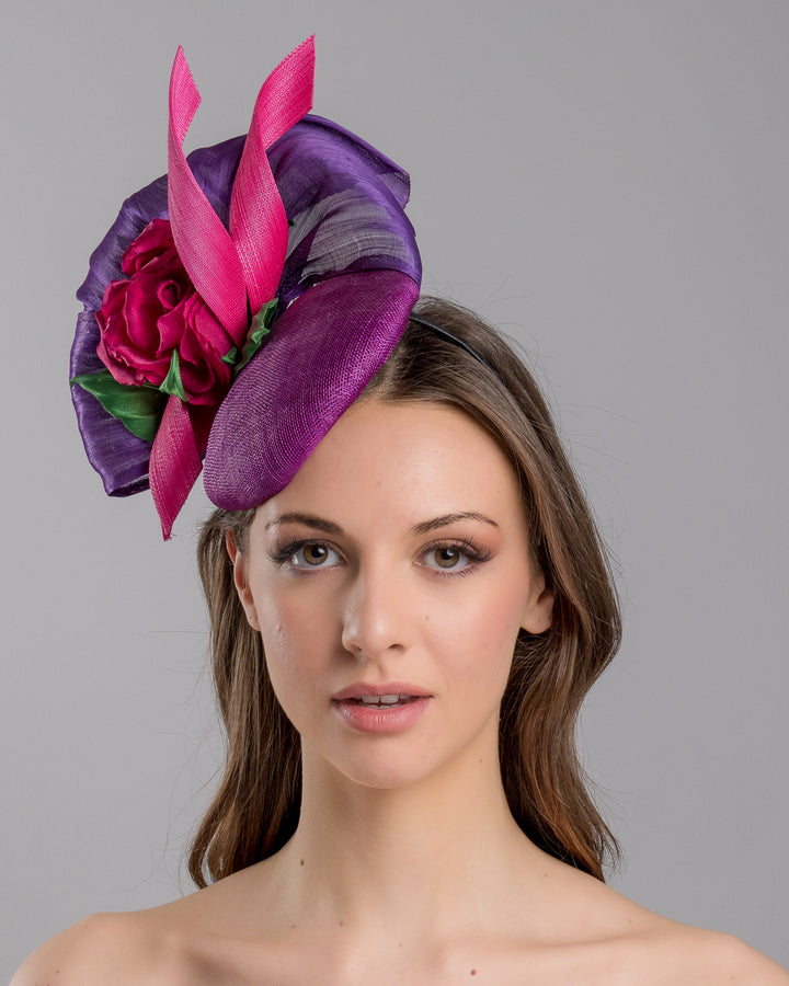 The model is showcasing the front side close up of the purple sinamay button shaped fascinator decorated with curved silk abaca fabric curving like a sea shell, two buntal swirls and a large pink silk rose, exemplifying the harmony of different shades of color. Designed and made by Merve Bayindir Millinery.
