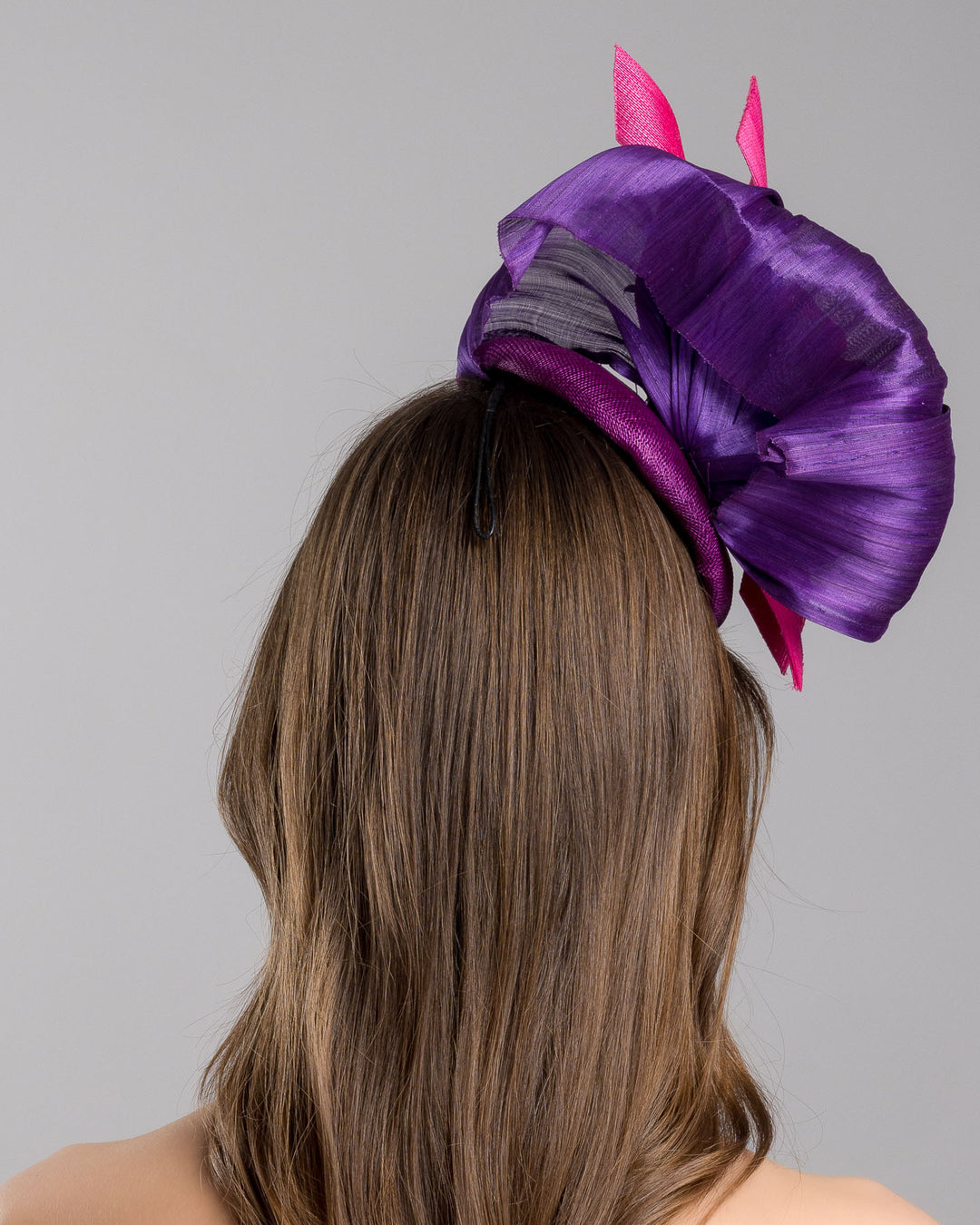 The model is showcasing the back side close up of the purple sinamay button shaped fascinator decorated with curved silk abaca fabric curving like a sea shell, two buntal swirls and a large pink silk rose, exemplifying the harmony of different shades of color. Designed and made by Merve Bayindir Millinery.
