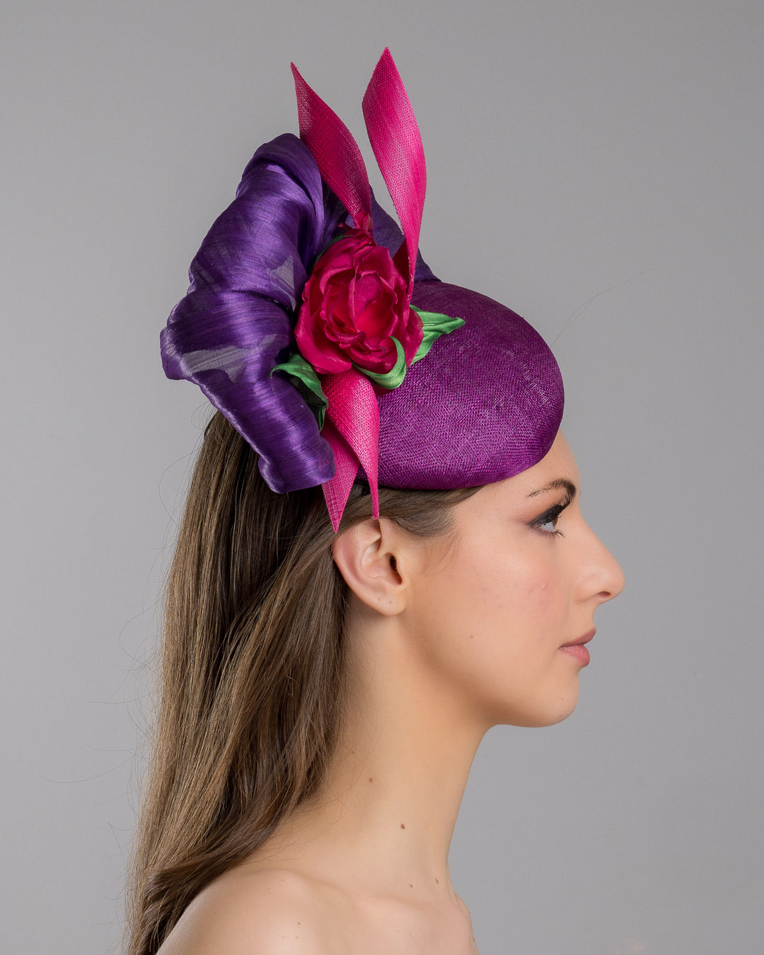 The model is showcasing the right side close up of the purple sinamay button shaped fascinator decorated with curved silk abaca fabric curving like a sea shell, two buntal swirls and a large pink silk rose, exemplifying the harmony of different shades of color. Designed and made by Merve Bayindir Millinery.

