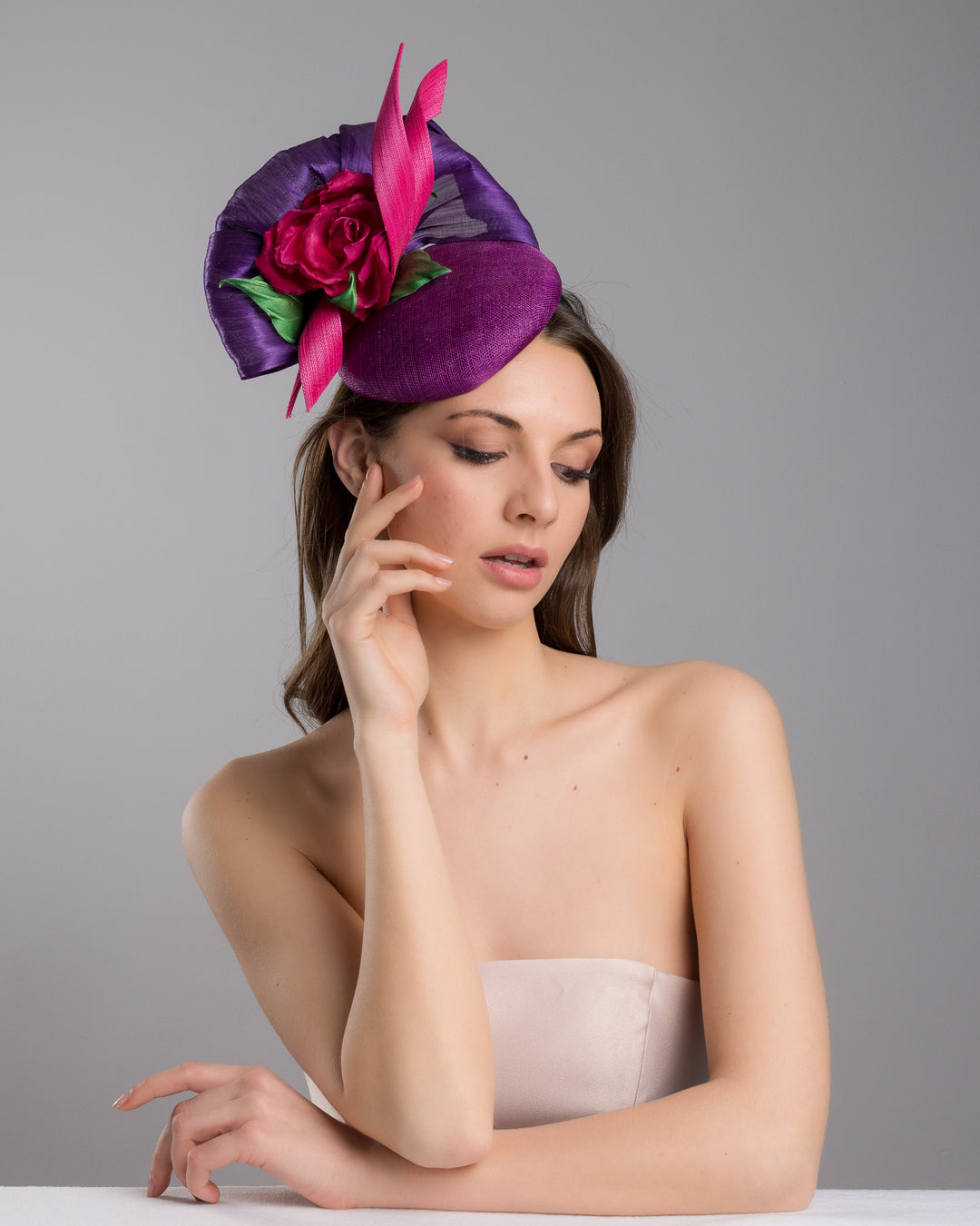 The female model is posing, wearing Tanner, the purple sinamay button shaped fascinator decorated with curved silk abaca fabric curving like a sea shell, two buntal swirls and a large pink silk rose, exemplifying the harmony of different shades of color. Designed and made by Merve Bayindir Millinery.
