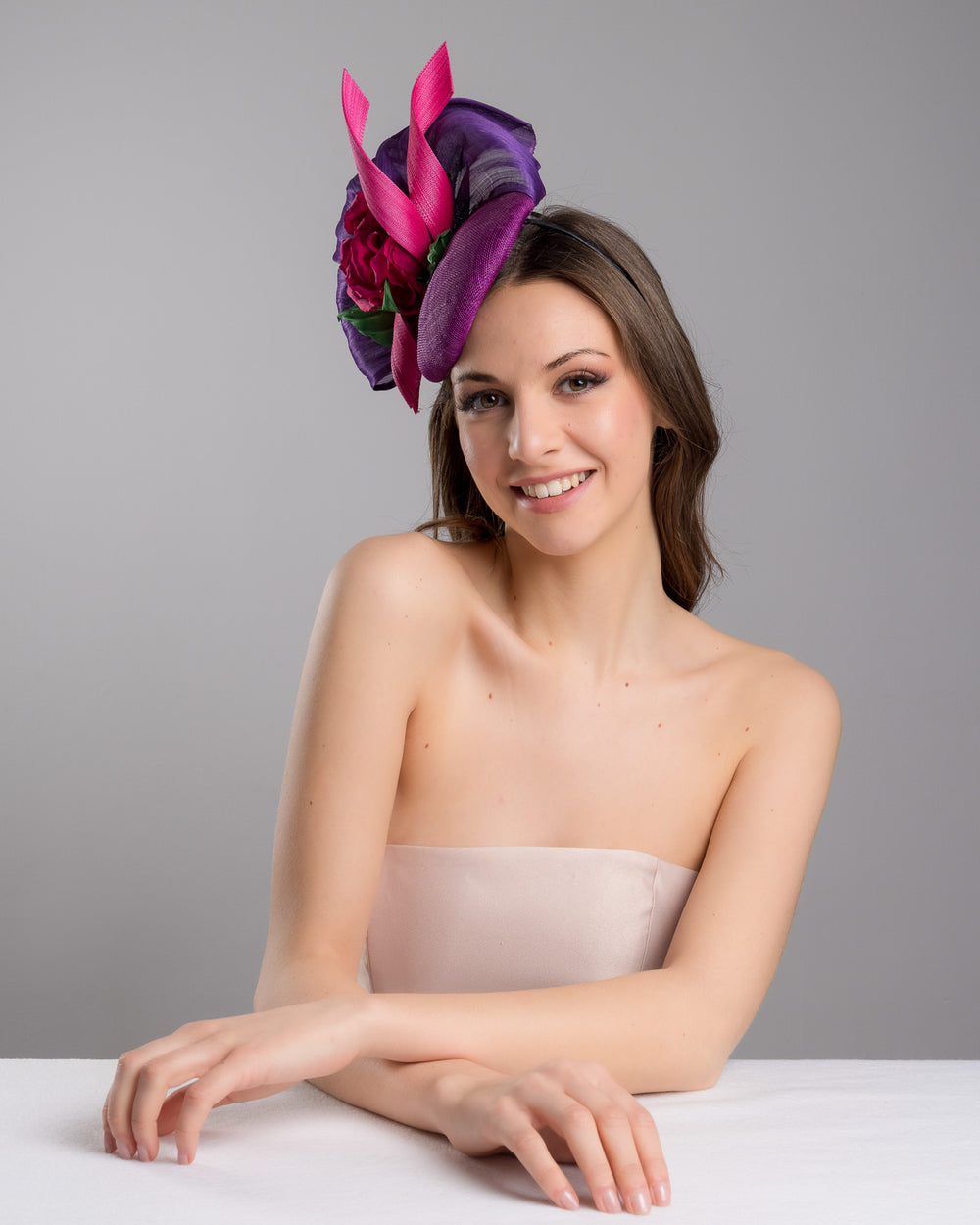 The female model is posing, wearing Tanner, the purple sinamay button shaped fascinator decorated with curved silk abaca fabric curving like a sea shell, two buntal swirls and a large pink silk rose, exemplifying the harmony of different shades of color. Designed and made by Merve Bayindir Millinery.
