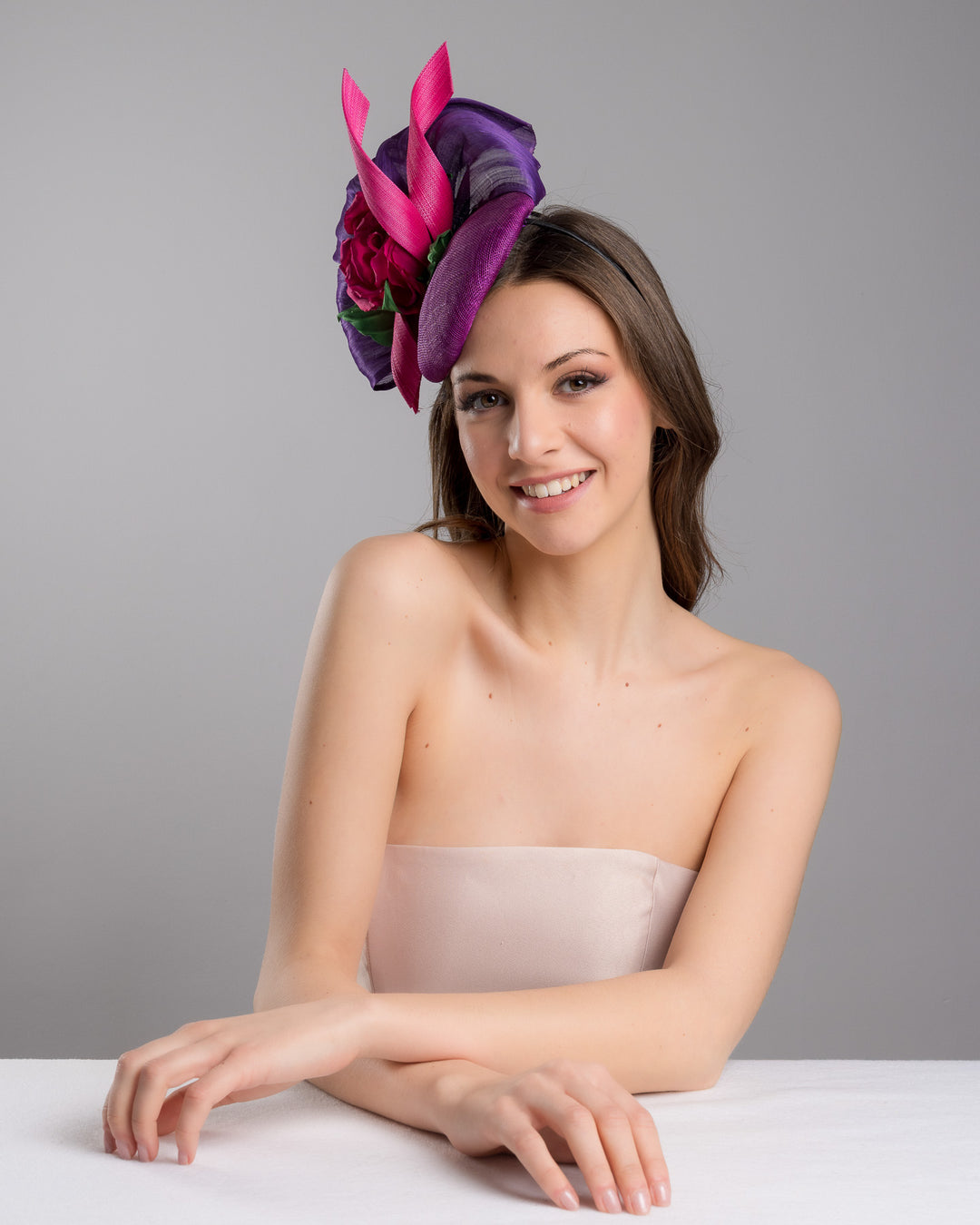 The female model is posing, wearing Tanner, the purple sinamay button shaped fascinator decorated with curved silk abaca fabric curving like a sea shell, two buntal swirls and a large pink silk rose, exemplifying the harmony of different shades of color. Designed and made by Merve Bayindir Millinery.
