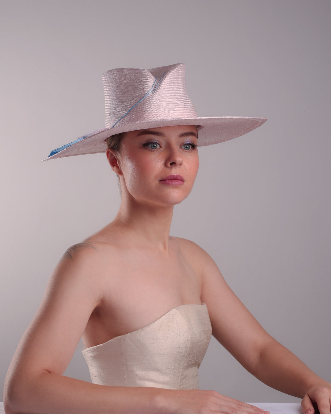 Model is showcasing the bottom front angle view a pink parasisal, wide flat brimmed hat with high curved crown, decorated with layered pink on top of a blue silk tassels.