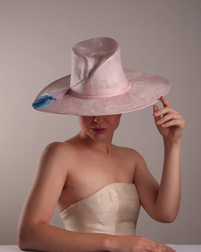 Model is showcasing the top angle view a pink parasisal, wide flat brimmed hat with high curved crown, decorated with layered pink on top of a blue silk tassels.