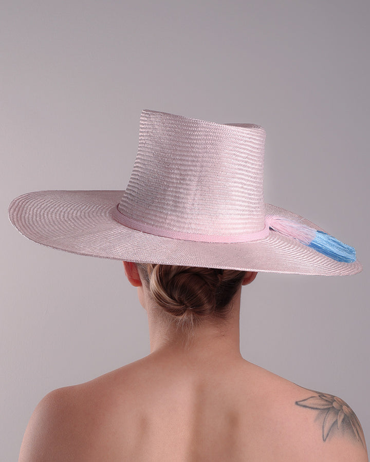 Model is showcasing the back of a pink parasisal, wide flat brimmed hat with high curved crown, decorated with layered pink on top of a blue silk tassels.