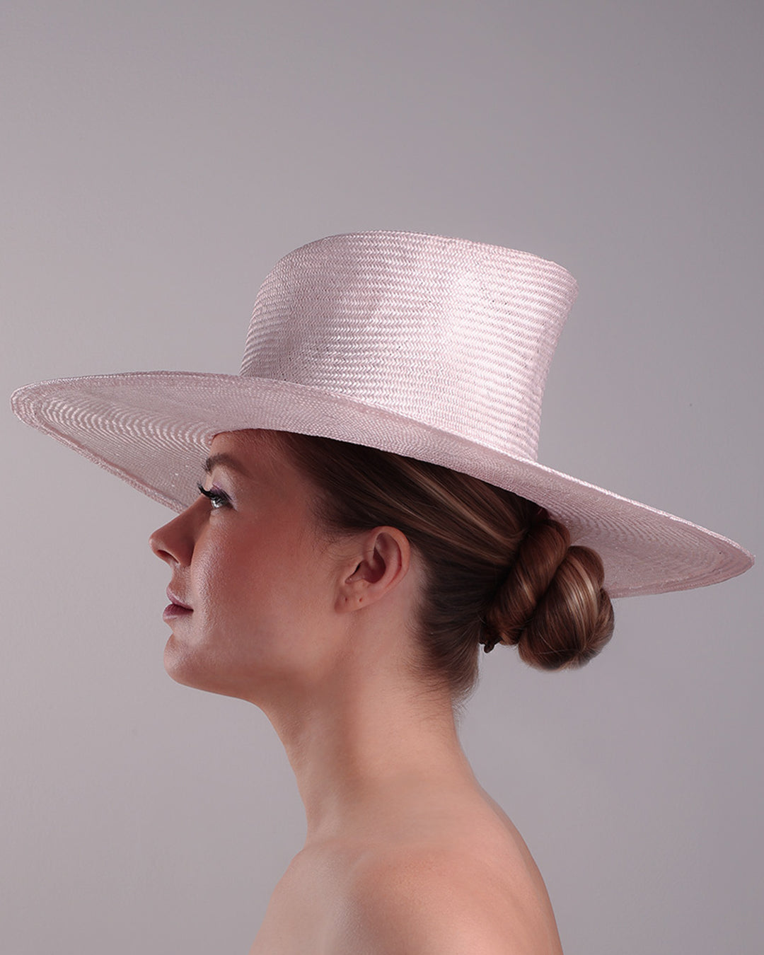 Model is showcasing the left side of a pink parasisal, wide flat brimmed hat with high curved crown, decorated with layered pink on top of a blue silk tassels.
