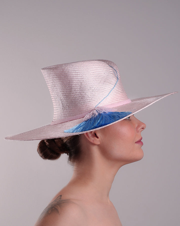 Model is showcasing the right side of a pink parasisal, wide flat brimmed hat with high curved crown, decorated with layered pink on top of a blue silk tassels.