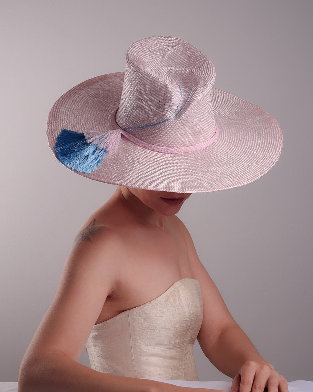 Model is showcasing the full top angle view a pink parasisal, wide flat brimmed hat with high curved crown, decorated with layered pink on top of a blue silk tassels.