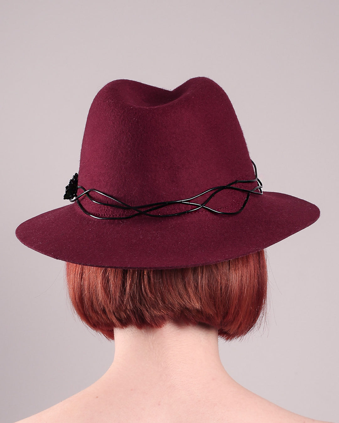 The model showcases the back of the Sylph fedora hat, a refined statement in deep burgundy felt, structured with clean, classic lines and softened by an ethereal twist: a fluid black wire detail weaves around the crown like delicate ink on parchment, with a crystal-embellished black metal swan on the left side. The embellishment feels both organic and intentional—a subtle nod to surrealism, grounded in wearable form.  Designed by Merve Bayindir.