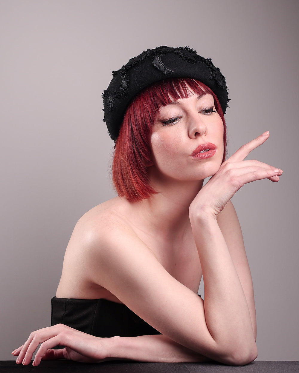 The model is showcasing the front below right angle of the Tilda pillbox beret hat. Made from structured black wool felt, the rounded dome-like silhouette is embroidered with a raised abstract motif in tonal thread, creating a sense of depth without distraction. Designed by Merve Bayindir.