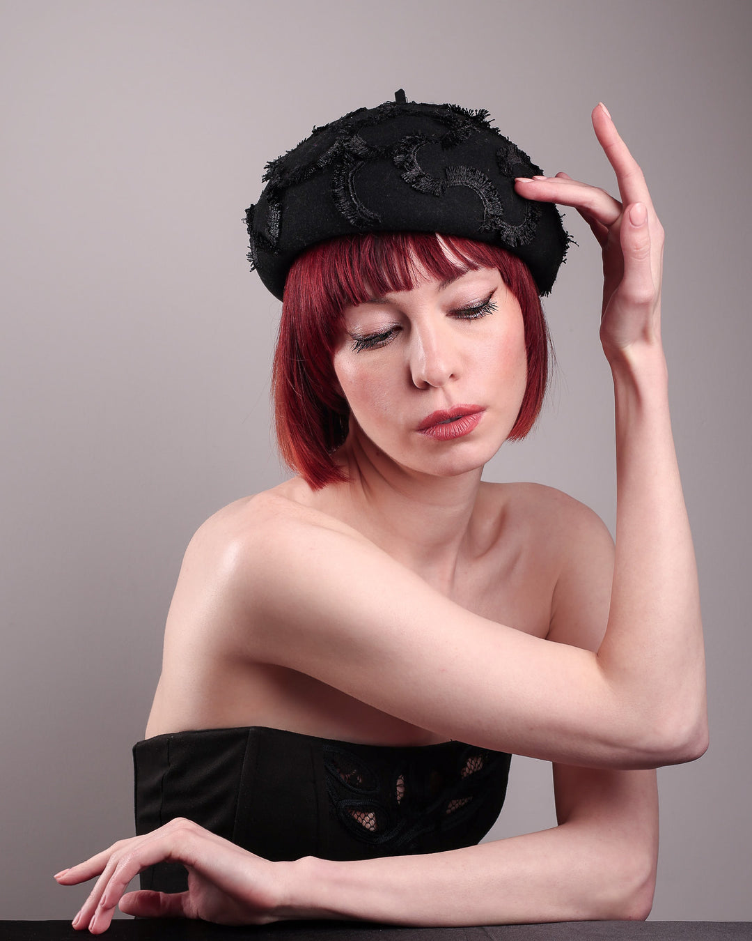 The model is showcasing the front side of the Tilda pillbox beret hat. Made from structured black wool felt, the rounded dome-like silhouette is embroidered with a raised abstract motif in tonal thread, creating a sense of depth without distraction. Designed by Merve Bayindir.