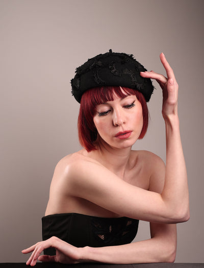 The model is showcasing the front side of the Tilda pillbox beret hat. Made from structured black wool felt, the rounded dome-like silhouette is embroidered with a raised abstract motif in tonal thread, creating a sense of depth without distraction. Designed by Merve Bayindir.