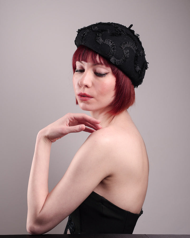 The model is showcasing the left front angle of the Tilda pillbox beret hat. Made from structured black wool felt, the rounded dome-like silhouette is embroidered with a raised abstract motif in tonal thread, creating a sense of depth without distraction. Designed by Merve Bayindir.