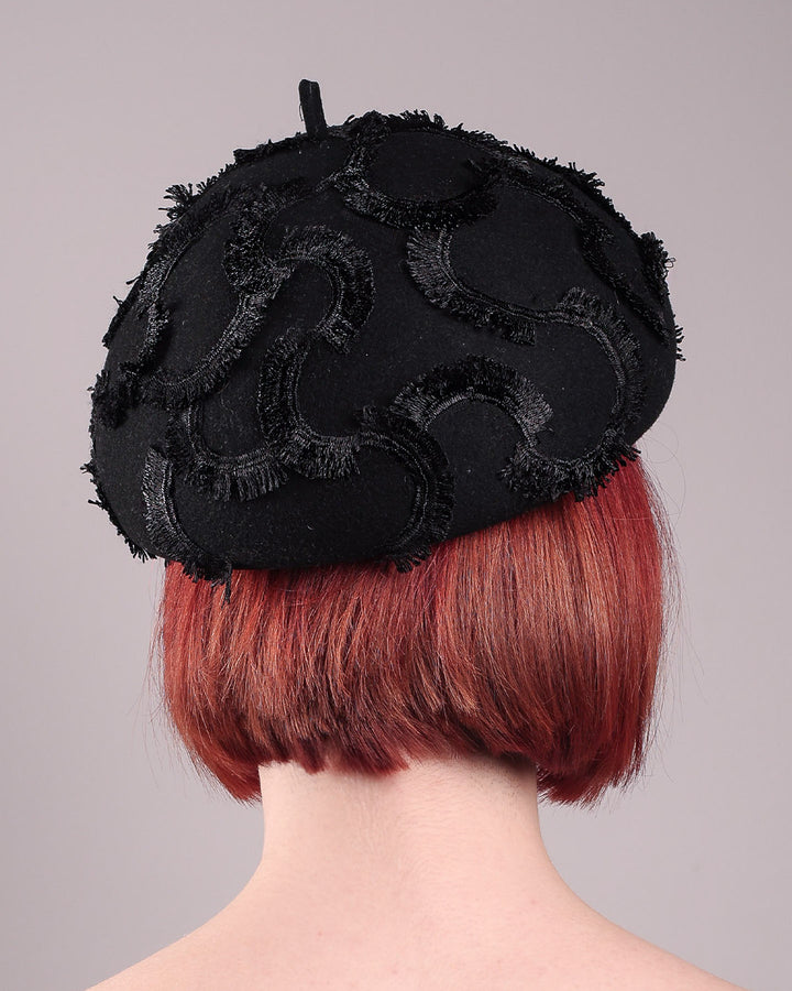 The model is showcasing the back of the Tilda pillbox beret hat. Made from structured black wool felt, the rounded dome-like silhouette is embroidered with a raised abstract motif in tonal thread, creating a sense of depth without distraction. Designed by Merve Bayindir.