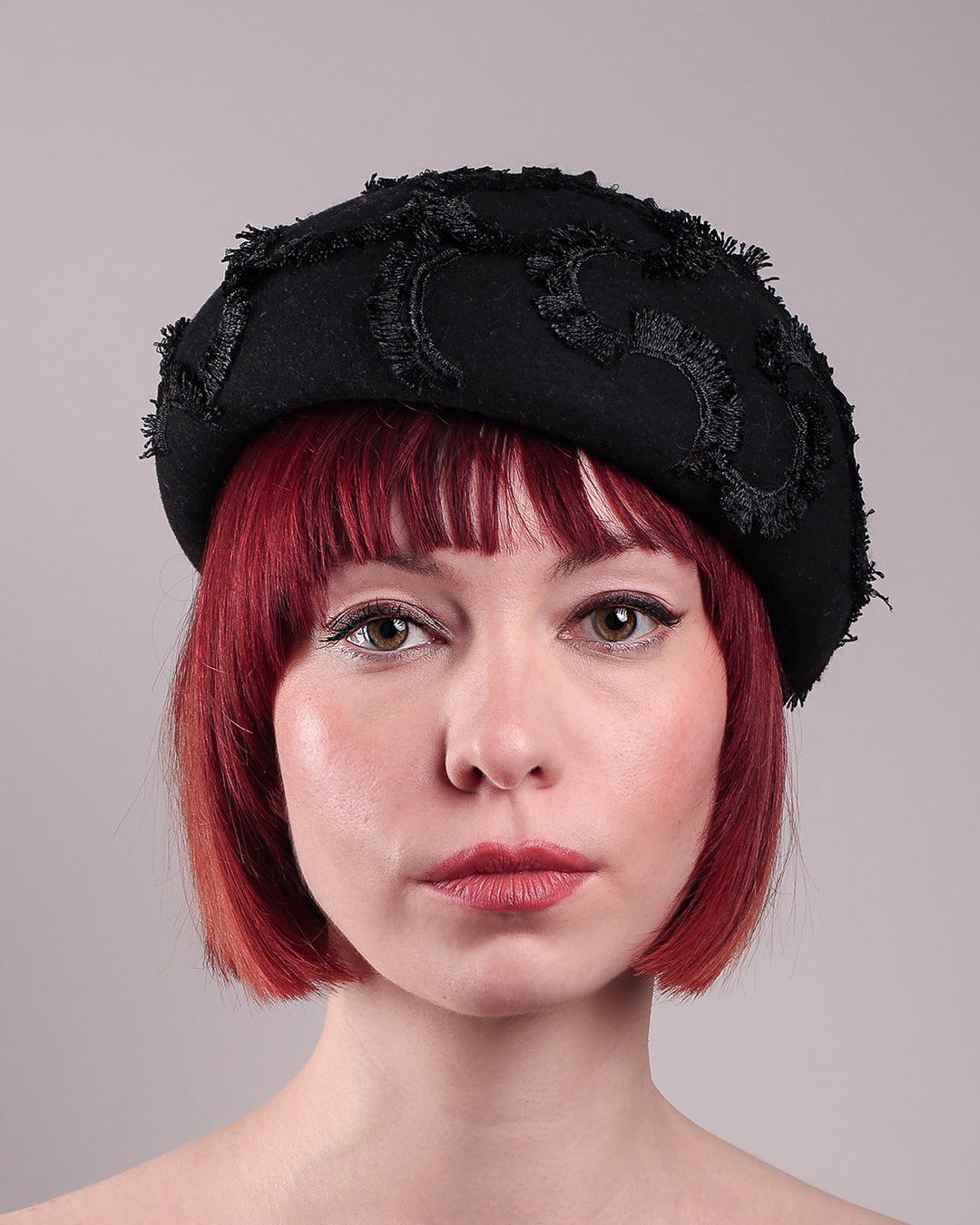 The model is showcasing the front close up of the Tilda pillbox beret hat. Made from structured black wool felt, the rounded dome-like silhouette is embroidered with a raised abstract motif in tonal thread, creating a sense of depth without distraction. Designed by Merve Bayindir.
