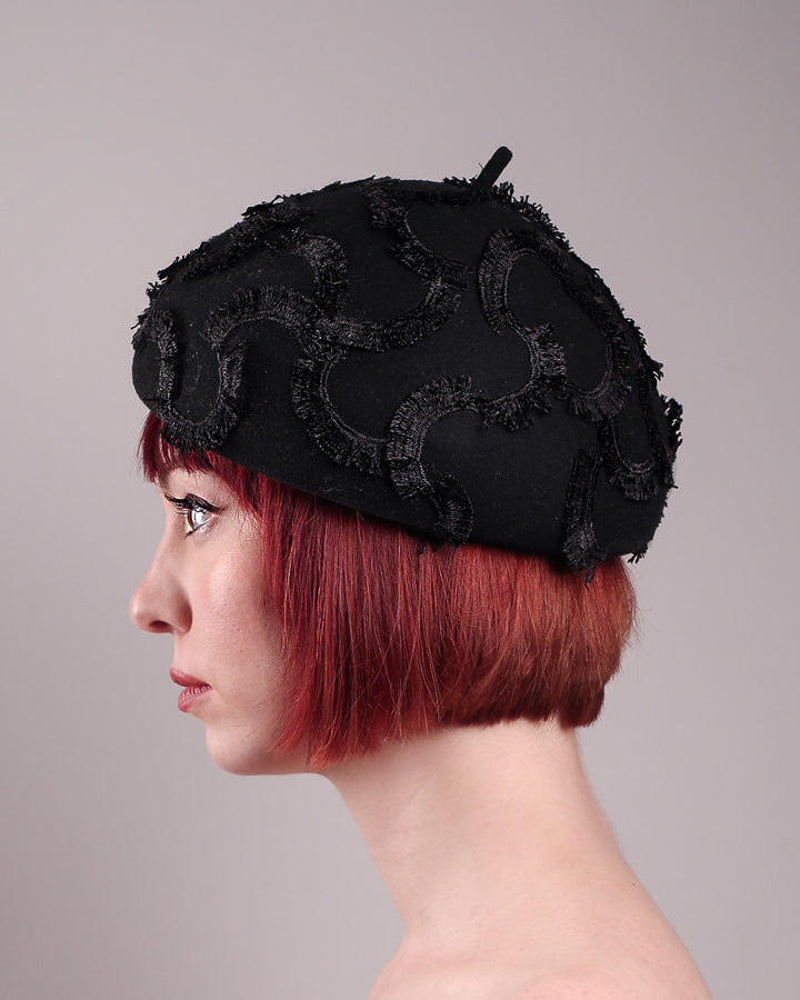 The model is showcasing the left side close up of the Tilda pillbox beret hat. Made from structured black wool felt, the rounded dome-like silhouette is embroidered with a raised abstract motif in tonal thread, creating a sense of depth without distraction. Designed by Merve Bayindir.
