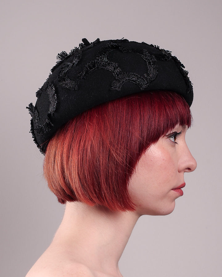 The model is showcasing the right side close up of the Tilda pillbox beret hat. Made from structured black wool felt, the rounded dome-like silhouette is embroidered with a raised abstract motif in tonal thread, creating a sense of depth without distraction. Designed by Merve Bayindir.