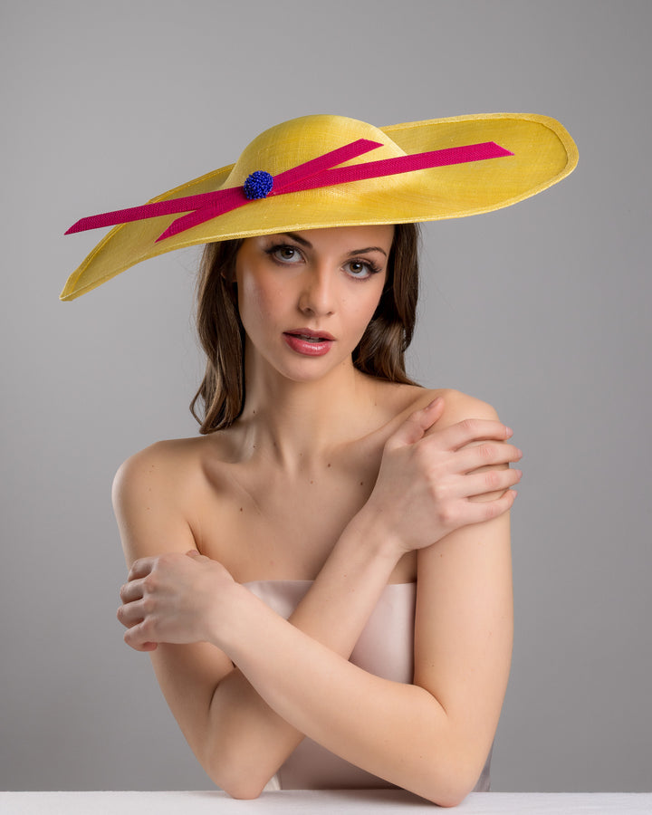 Female model posing wearing the Timber  wide brim asymmetric yellow hat tilted to the left. Decorated with two pink buntal stripes crossing the hat as well as each other, embellished the centre of straight ribbon is a dark blue bead bead cluster. Designed and made by Merve Bayindir millinery.
