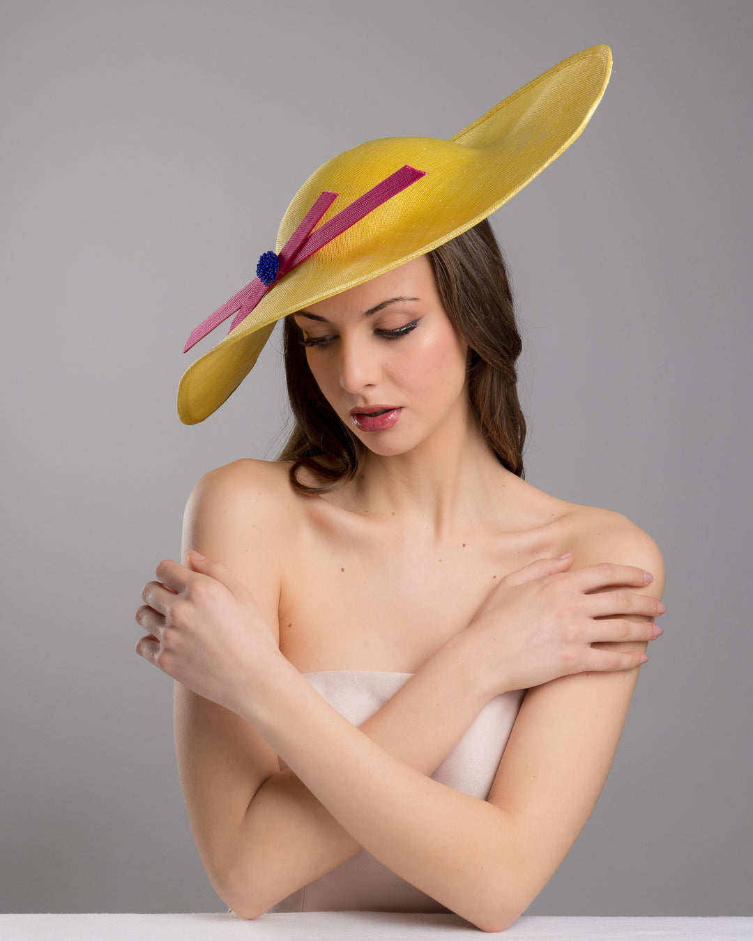 Female model posing wearing the Timber  wide brim asymmetric yellow hat tilted to the left. Decorated with two pink buntal stripes crossing the hat as well as each other, embellished the centre of straight ribbon is a dark blue bead bead cluster. Designed and made by Merve Bayindir millinery.