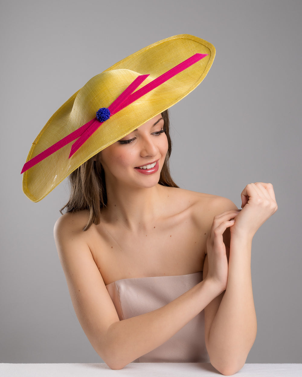 Female model posing wearing the Timber  wide brim asymmetric yellow hat tilted to the left. Decorated with two pink buntal stripes crossing the hat as well as each other, embellished the centre of straight ribbon is a dark blue bead bead cluster. Designed and made by Merve Bayindir millinery.
