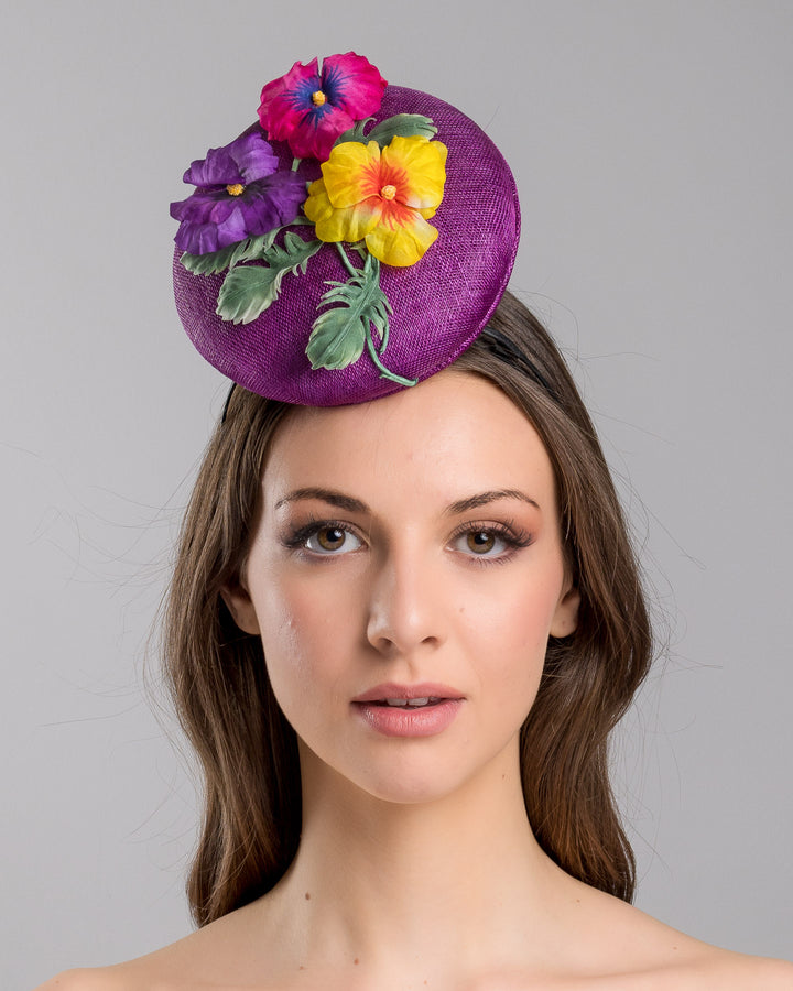 The model is showcasing the front side close up of the purple button shape fascinator, that sirs on the centre of the head, the fascinator is decorated with a purplue and a pink and a yellow violet bouquet with stems and leaves. Designed and made by Merve Bayindir Millinery