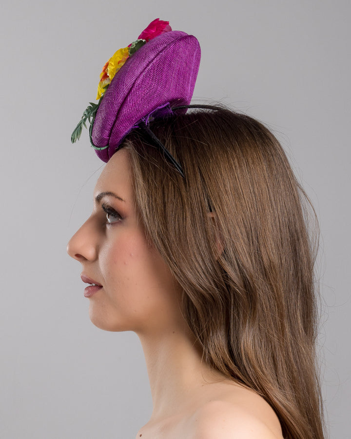 The model is showcasing the left side close up of the purple button shape fascinator, that sirs on the centre of the head, the fascinator is decorated with a purplue and a pink and a yellow violet bouquet with stems and leaves. Designed and made by Merve Bayindir Millinery