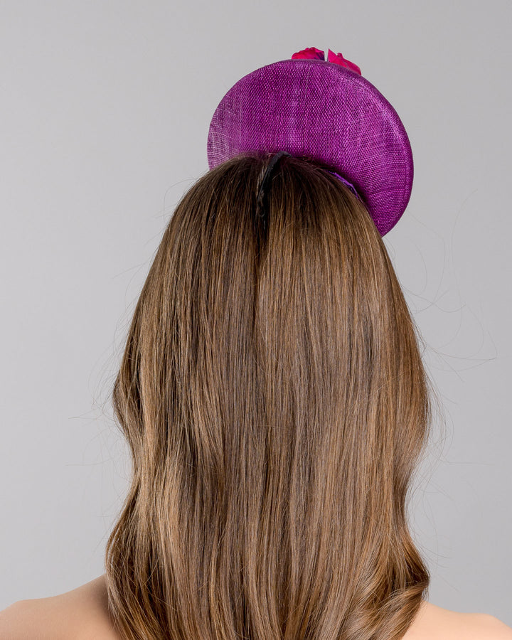 The model is showcasing the back side close up of the purple button shape fascinator, that sirs on the centre of the head, the fascinator is decorated with a purplue and a pink and a yellow violet bouquet with stems and leaves. Designed and made by Merve Bayindir Millinery