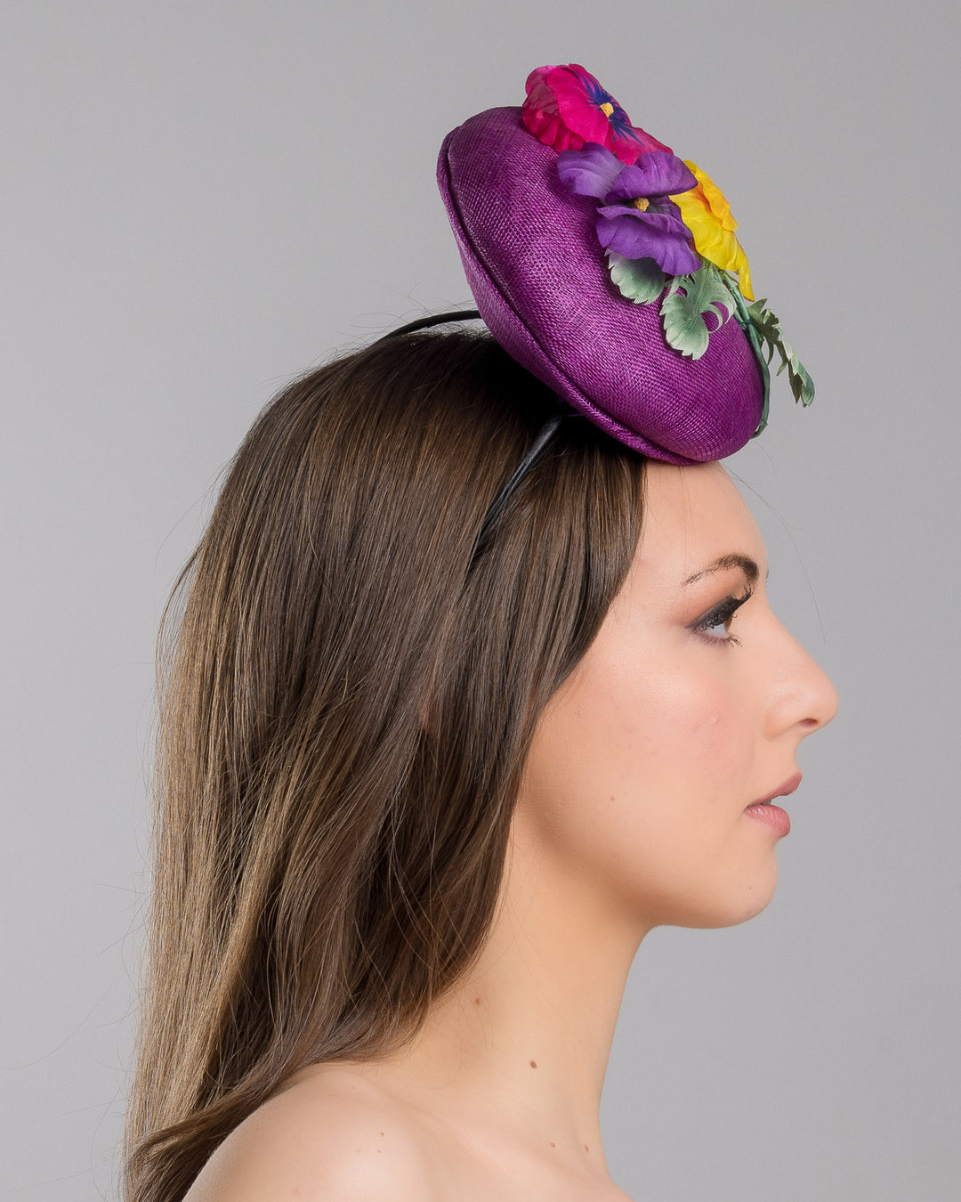 The model is showcasing the right side close up of the purple button shape fascinator, that sirs on the centre of the head, the fascinator is decorated with a purplue and a pink and a yellow violet bouquet with stems and leaves. Designed and made by Merve Bayindir Millinery