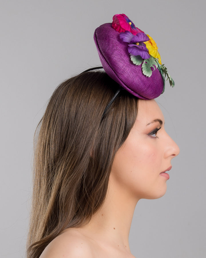 The model is showcasing the right side close up of the purple button shape fascinator, that sirs on the centre of the head, the fascinator is decorated with a purplue and a pink and a yellow violet bouquet with stems and leaves. Designed and made by Merve Bayindir Millinery