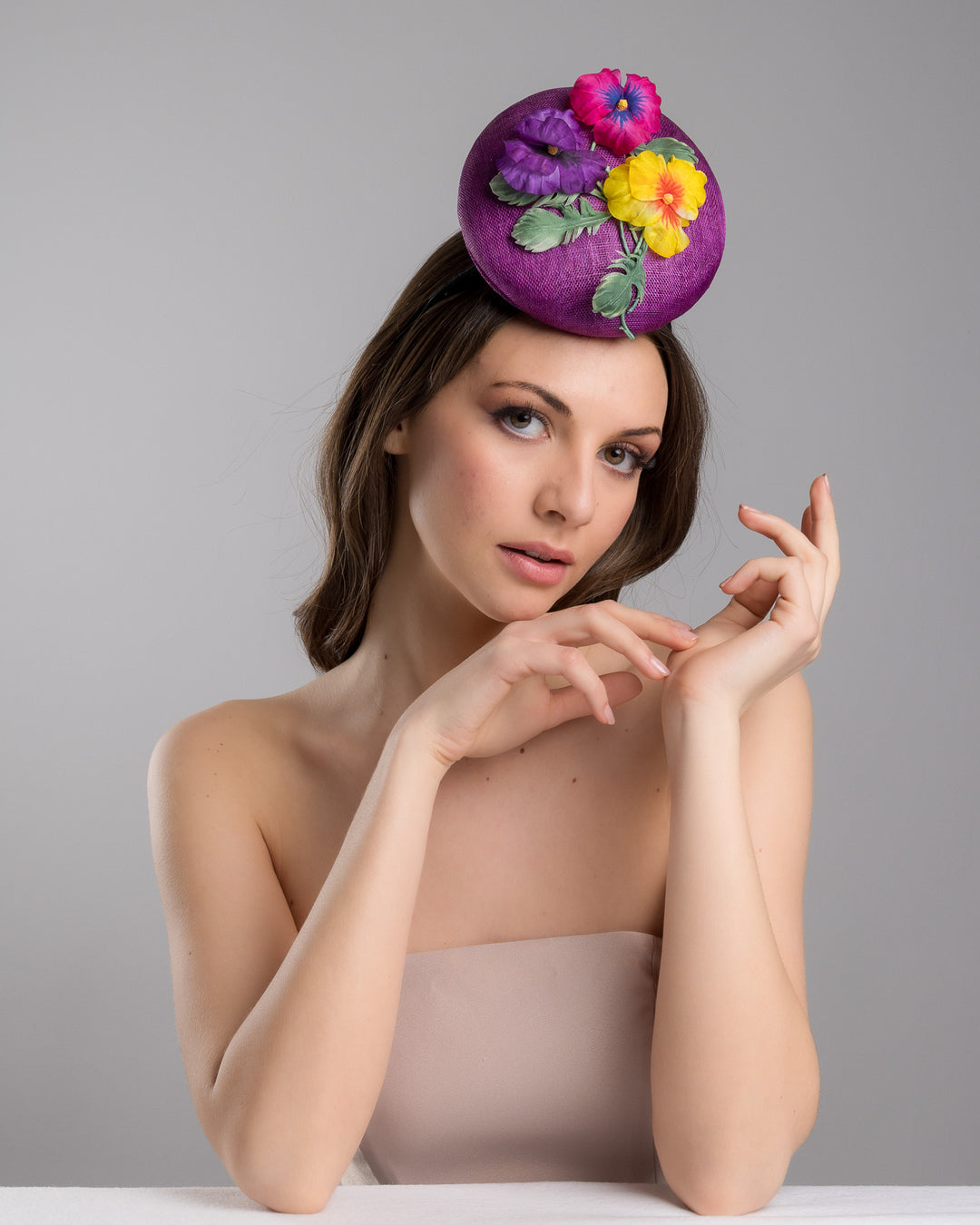 The female model is posing, wearing Tommie, the purple button shape fascinator, that sirs on the centre of the head, the fascinator is decorated with a purplue and a pink and a yellow violet bouquet with stems and leaves. Designed and made by Merve Bayindir Millinery