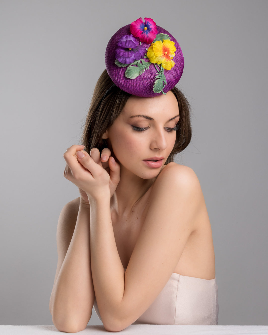 The female model is posing, wearing Tommie, the purple button shape fascinator, that sirs on the centre of the head, the fascinator is decorated with a purplue and a pink and a yellow violet bouquet with stems and leaves. Designed and made by Merve Bayindir Millinery