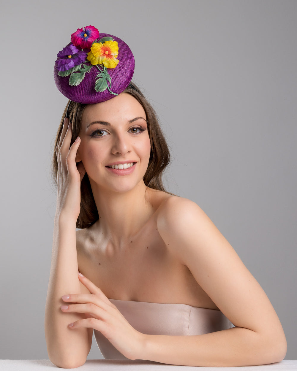 The female model is posing, wearing Tommie, the purple button shape fascinator, that sirs on the centre of the head, the fascinator is decorated with a purplue and a pink and a yellow violet bouquet with stems and leaves. Designed and made by Merve Bayindir Millinery