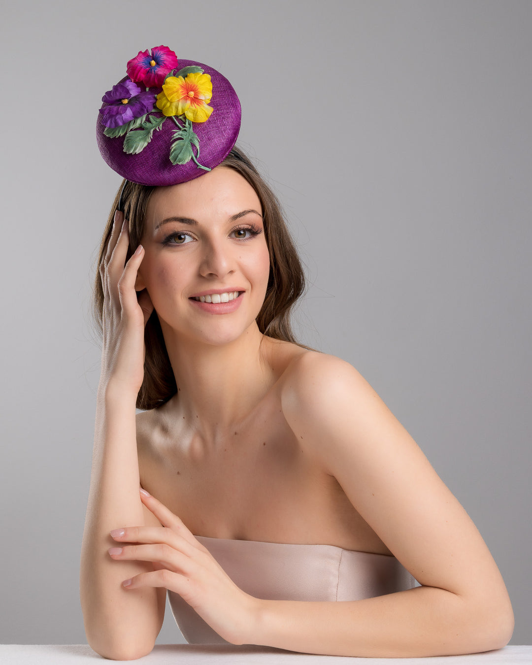 The female model is posing, wearing Tommie, the purple button shape fascinator, that sirs on the centre of the head, the fascinator is decorated with a purplue and a pink and a yellow violet bouquet with stems and leaves. Designed and made by Merve Bayindir Millinery