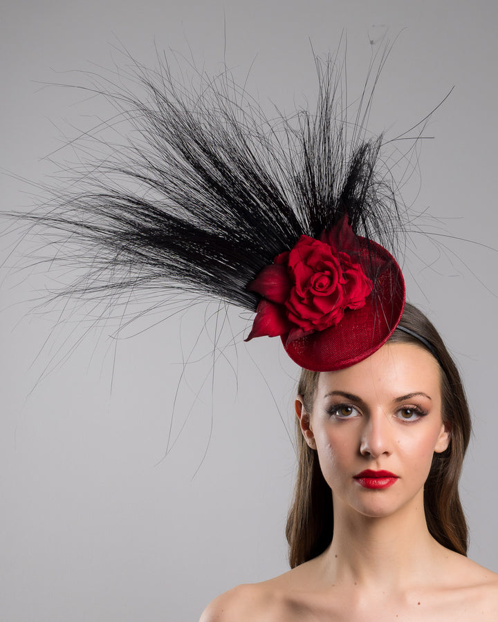 Model is showcasing the front side close up of mini red saucer fascinator, adorned with a red rose embellished with high rising black burnt peacock feathers. Designed and made by Merve Bayindir millinery.