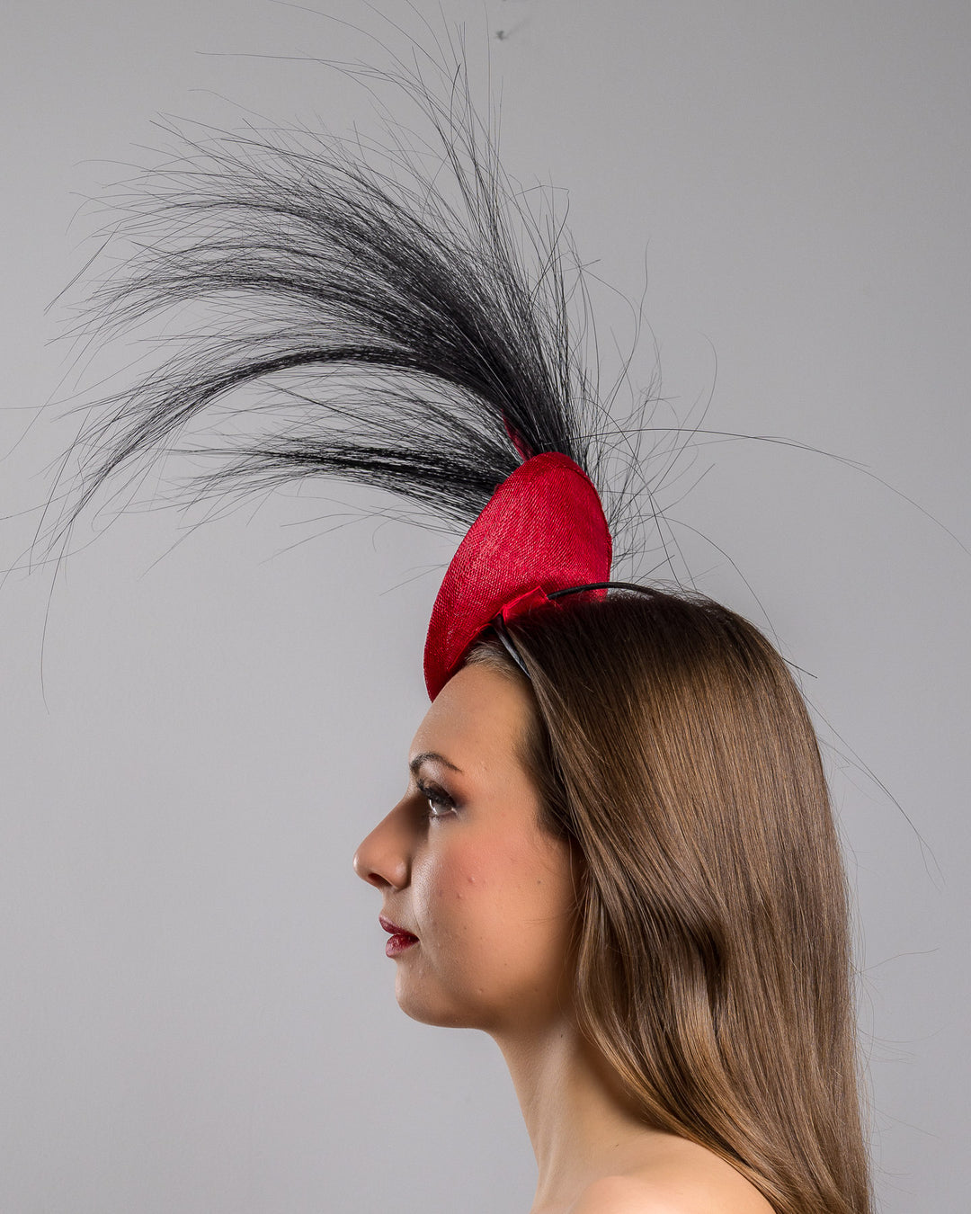 Model is showcasing the left side close up of mini red saucer fascinator, adorned with a red rose embellished with high rising black burnt peacock feathers. Designed and made by Merve Bayindir millinery.