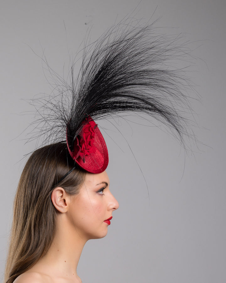 Model is showcasing the right side close up of mini red saucer fascinator, adorned with a red rose embellished with high rising black burnt peacock feathers. Designed and made by Merve Bayindir millinery.