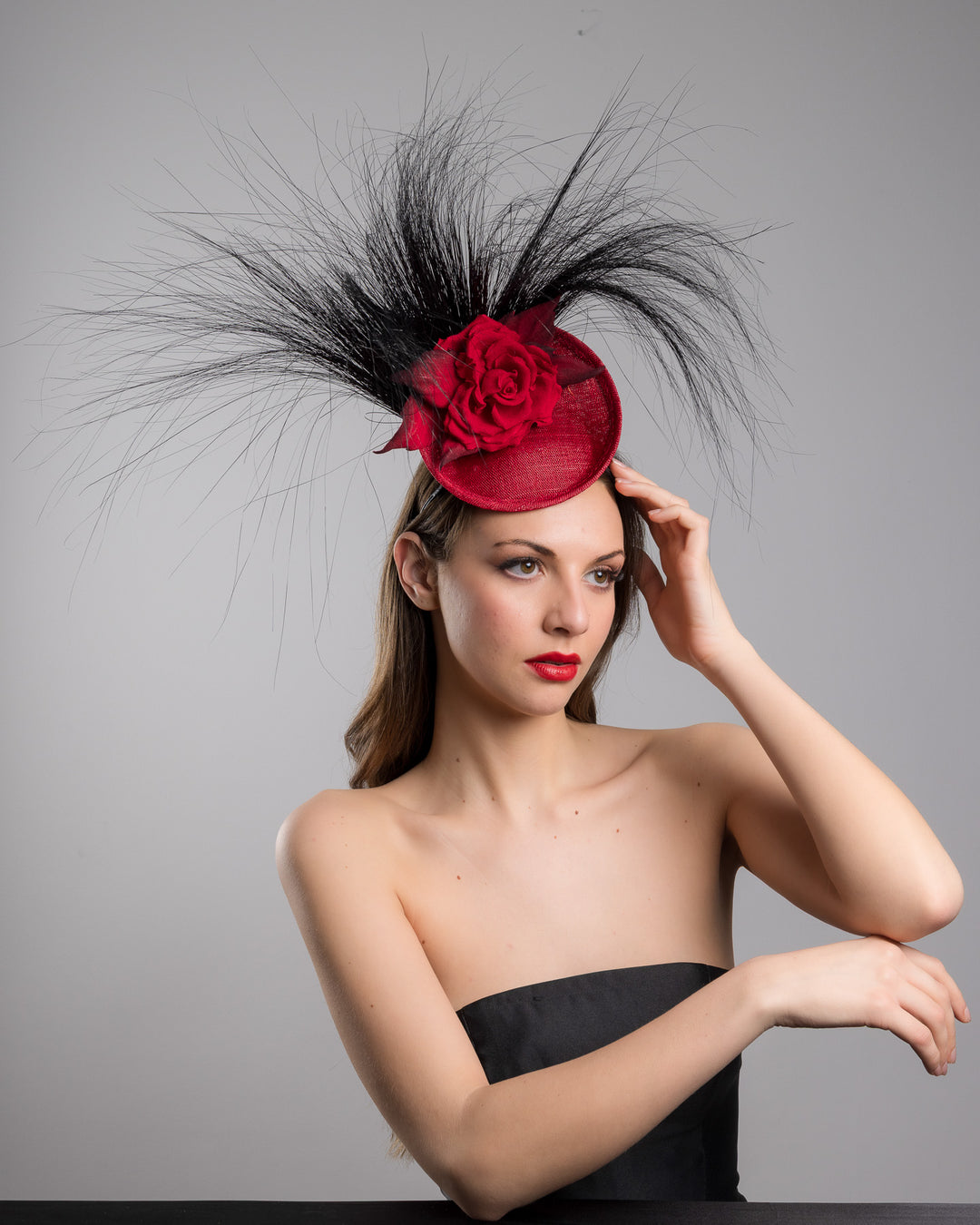 Female model is posing wearing the high fashion Trust mini red saucer fascinator, adorned with a red rose embellished with high rising black burnt peacock feathers. Designed and made by Merve Bayindir millinery.