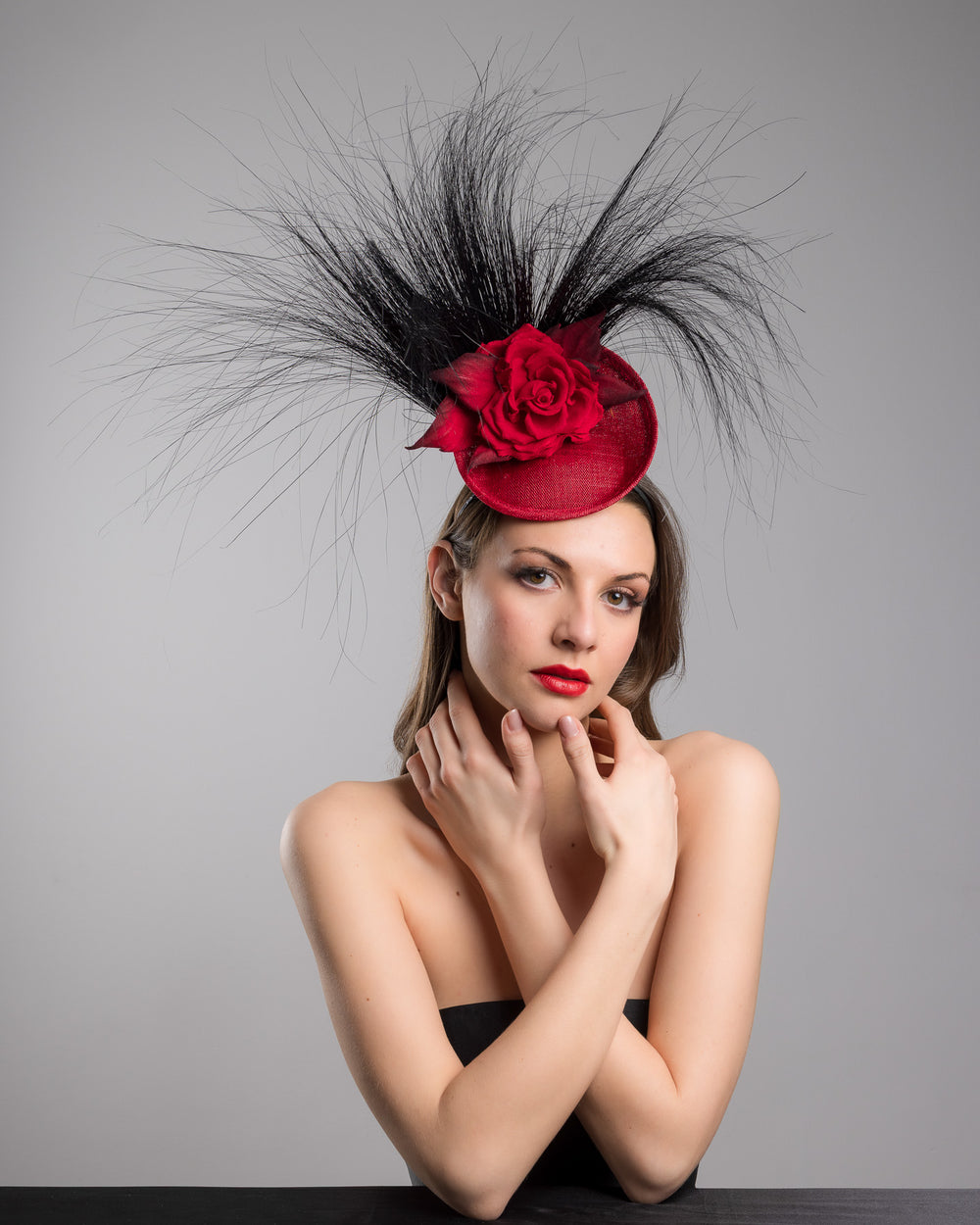 Female model is posing wearing the high fashion Trust mini red saucer fascinator, adorned with a red rose embellished with high rising black burnt peacock feathers. Designed and made by Merve Bayindir millinery.