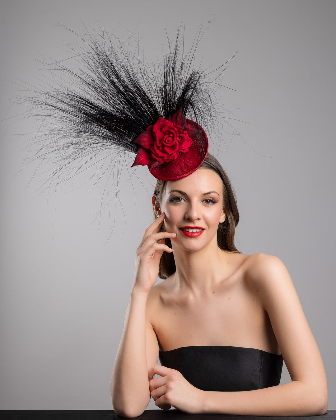 Female model is posing wearing the high fashion Trust mini red saucer fascinator, adorned with a red rose embellished with high rising black burnt peacock feathers. Designed and made by Merve Bayindir millinery.