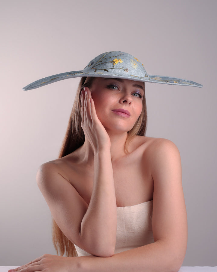 The model is showcasing the front of a blue asymetric whide brim hat, tilted to the right, decorated with pressed natural flowers creatinga almost 3D dimensional print affect on the hat. Fitted with a wired headband and additionally secured with elastic.

