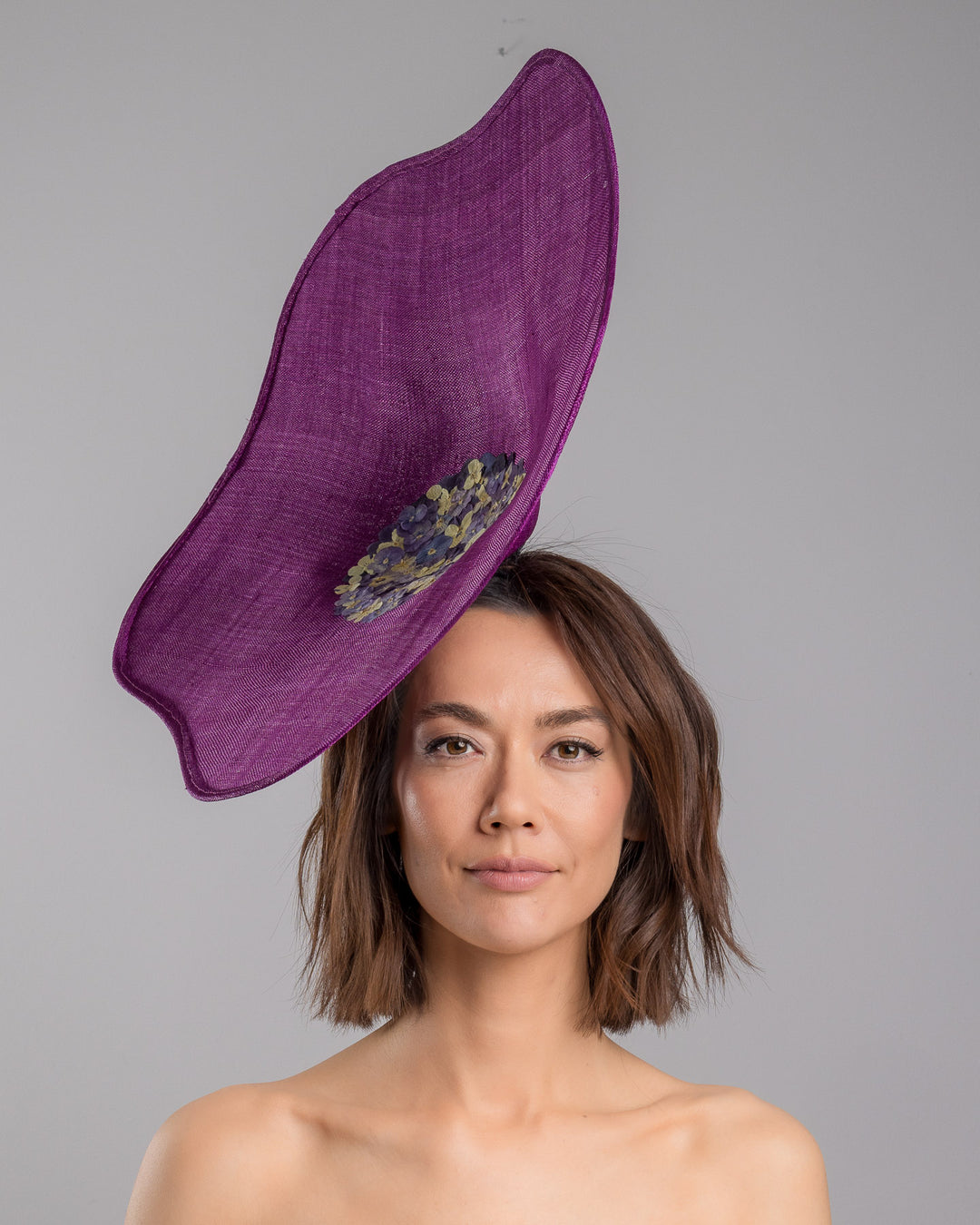 The model is showcasing the front side close up of the purple sinamay statement fascinator with an organically sculpted, flowing silhouette, featuring a central cluster of delicate yellow and violet flowers for a vibrant, artistic finish. Designed and made by Merve Bayindir Millinery.
