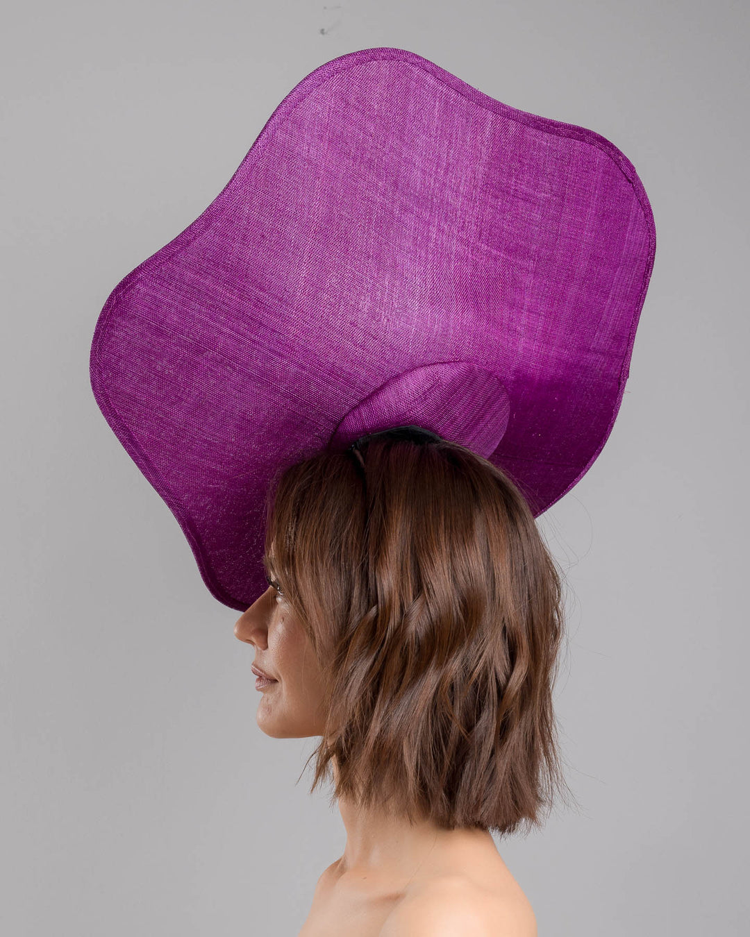 The model is showcasing the left side close up of the purple sinamay statement fascinator with an organically sculpted, flowing silhouette, featuring a central cluster of delicate yellow and violet flowers for a vibrant, artistic finish. Designed and made by Merve Bayindir Millinery.
