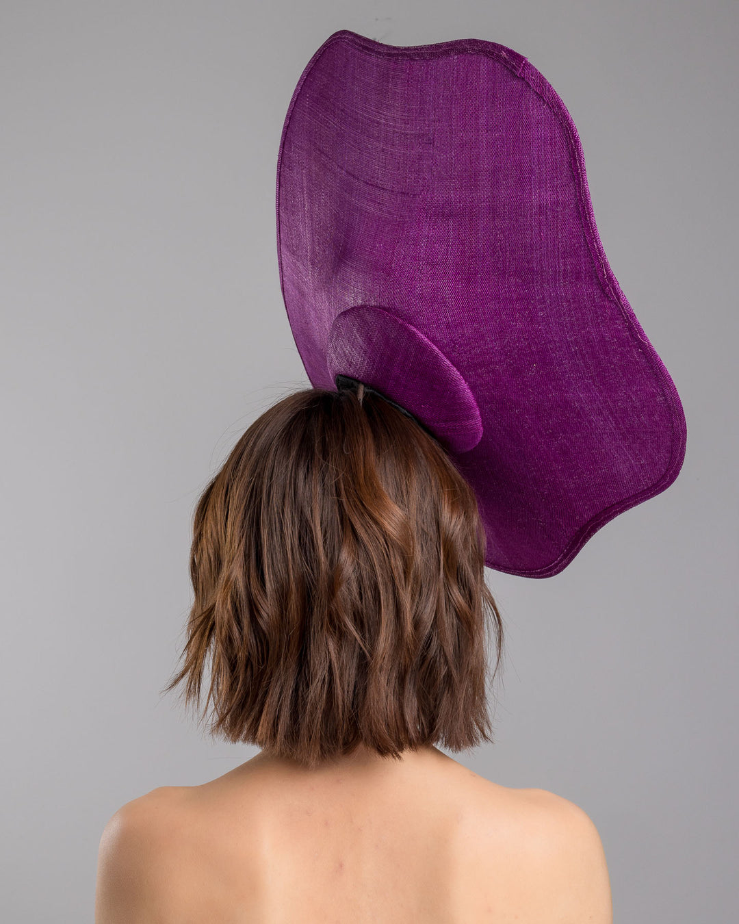 The model is showcasing the back side close up of the purple sinamay statement fascinator with an organically sculpted, flowing silhouette, featuring a central cluster of delicate yellow and violet flowers for a vibrant, artistic finish. Designed and made by Merve Bayindir Millinery.
