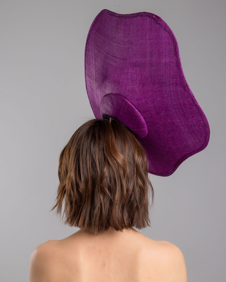 The model is showcasing the back side close up of the purple sinamay statement fascinator with an organically sculpted, flowing silhouette, featuring a central cluster of delicate yellow and violet flowers for a vibrant, artistic finish. Designed and made by Merve Bayindir Millinery.
