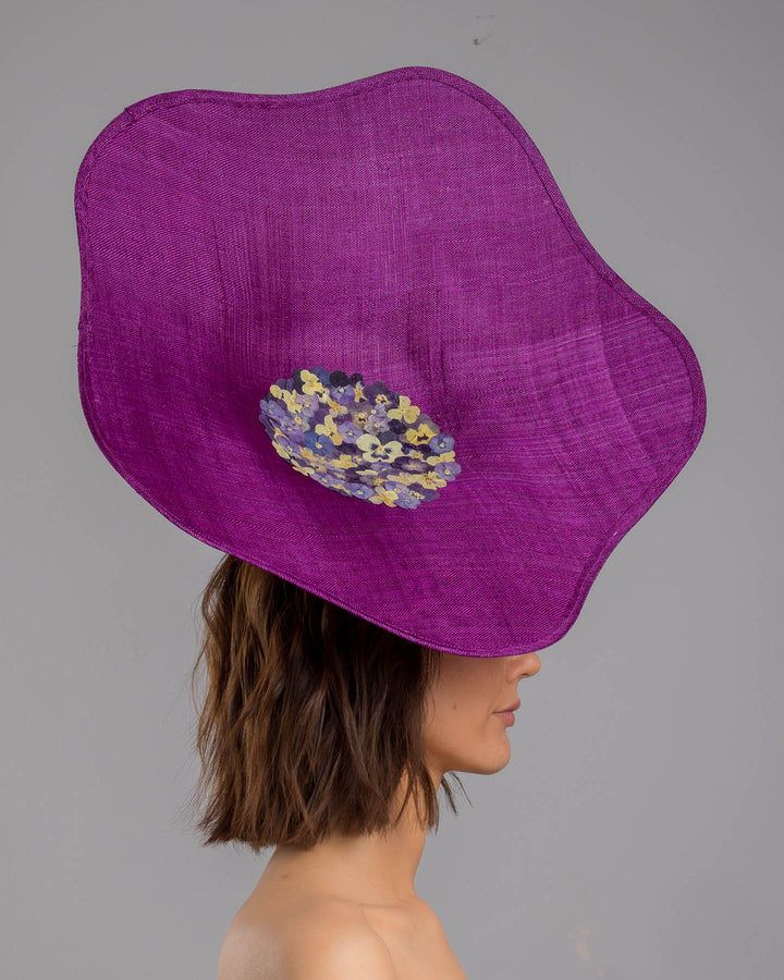 The model is showcasing the right side close up of the purple sinamay statement fascinator with an organically sculpted, flowing silhouette, featuring a central cluster of delicate yellow and violet flowers for a vibrant, artistic finish. Designed and made by Merve Bayindir Millinery.
