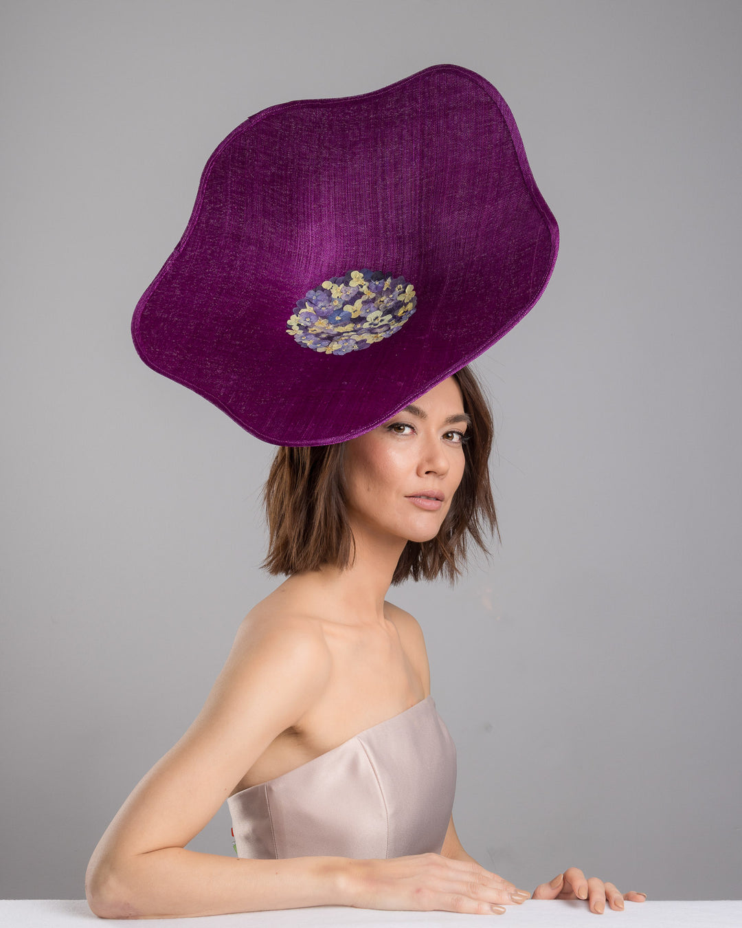 The female model is posing, wearing
Ulma, the purple sinamay statement fascinator with an organically sculpted, flowing silhouette, featuring a central cluster of delicate yellow and violet flowers for a vibrant, artistic finish. Designed and made by Merve Bayindir Millinery.